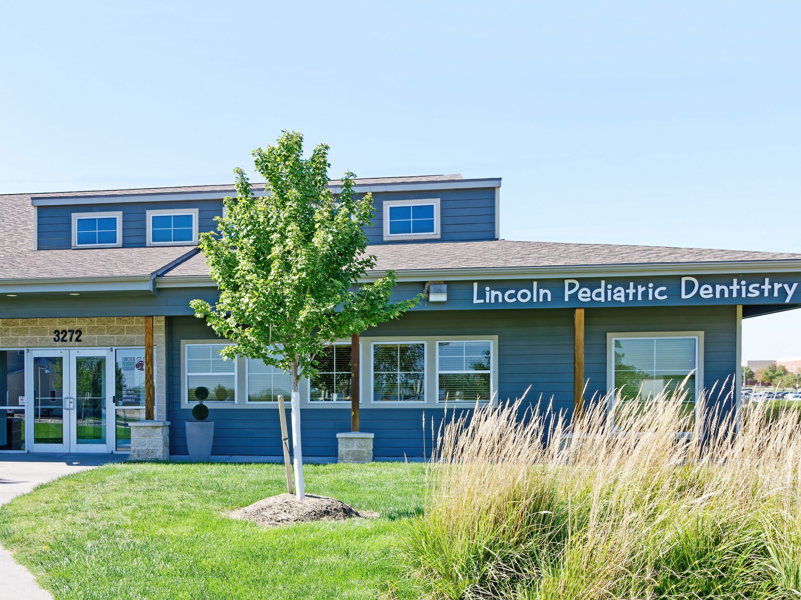 A modern blue building labeled "Lincoln Pediatric Dentistry" has large windows, a neatly trimmed lawn, and a small tree in front.
