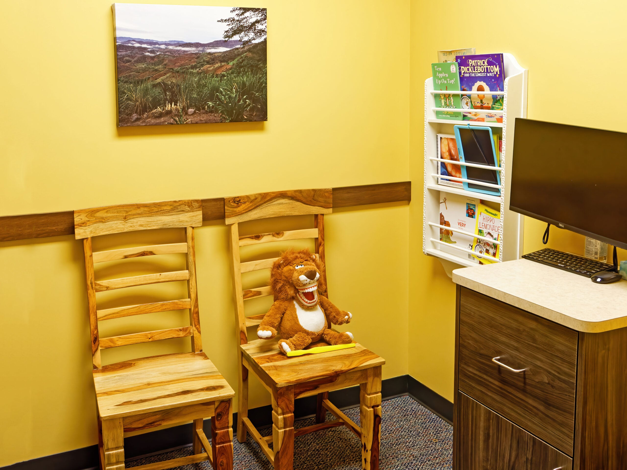A cozy corner with two wooden chairs, a plush lion toy, wall art, brochures, and a monitor in a yellow-painted room.