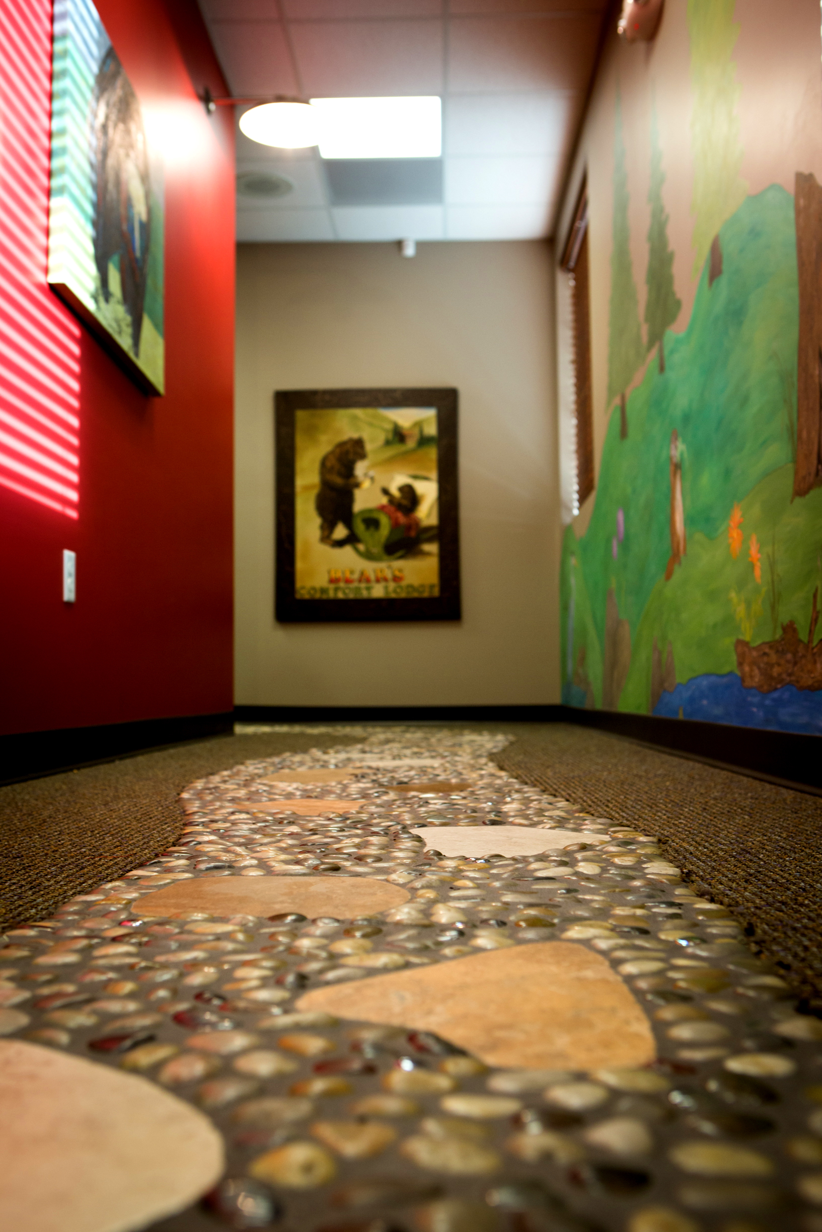 A hallway with stone-textured flooring, colorful murals, and bear artwork on the wall. Ceiling lights illuminate the vibrant red and green walls.