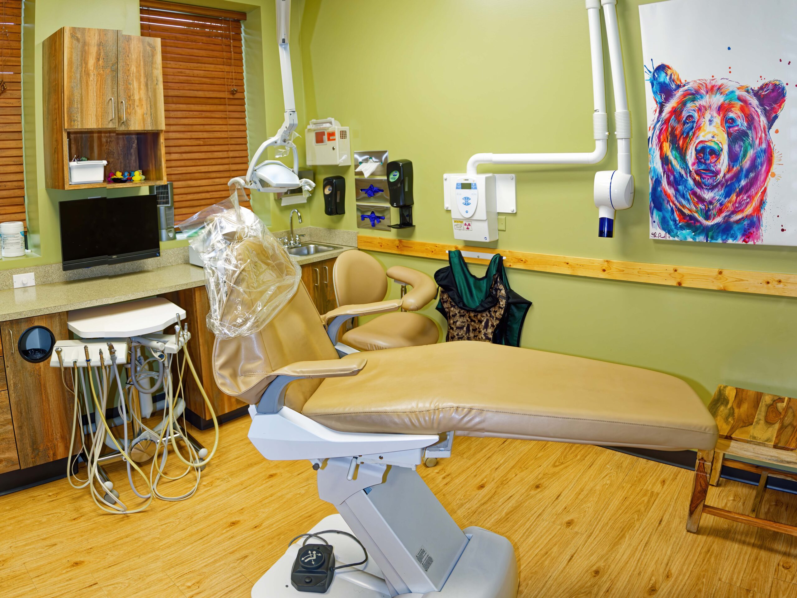 A modern dental office with a reclining chair, medical equipment, and a colorful bear painting on a green wall, creating a relaxed atmosphere.