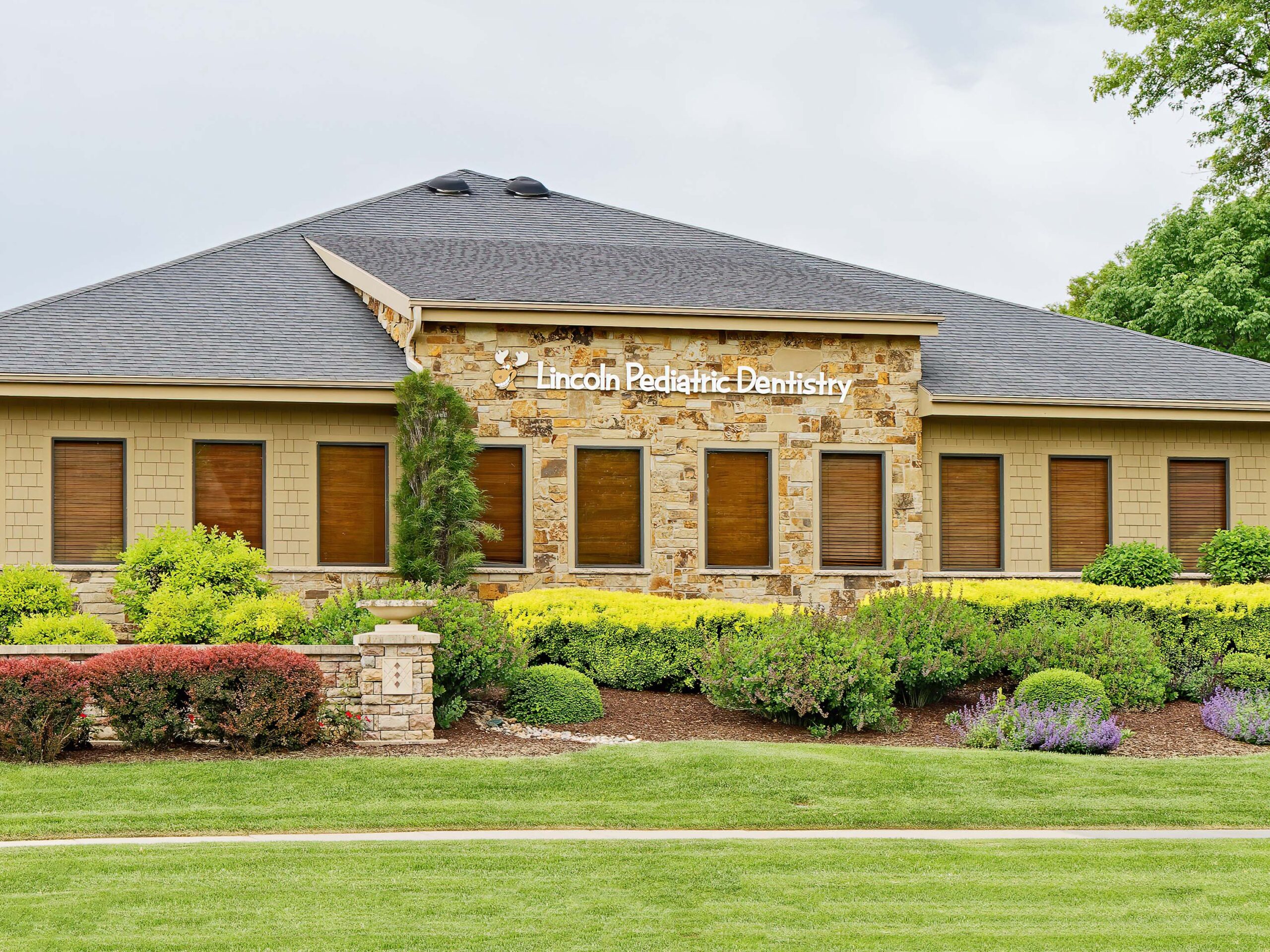 A stone building with a sign reading "Lincoln Pediatric Dentistry" is surrounded by landscaped greenery and shrubs under a partly cloudy sky.