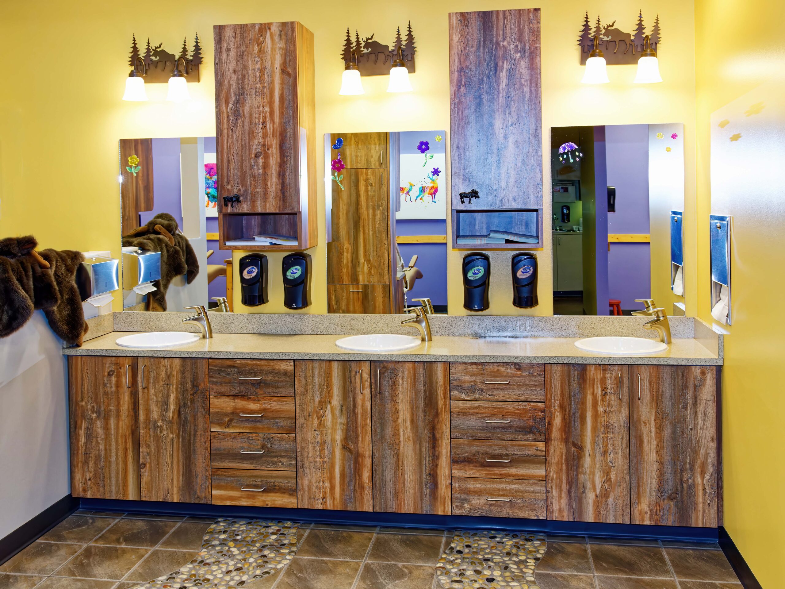 Bright, rustic bathroom with wooden cabinetry, dual sinks, three soap dispensers, and decorative moose motifs. Warm lighting enhances the yellow walls.
