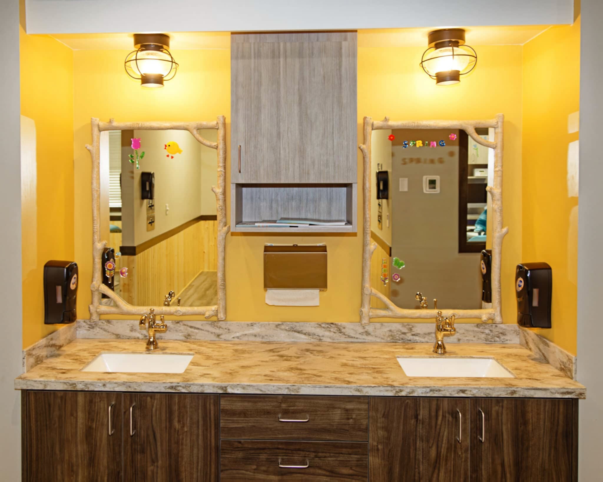 Bright yellow bathroom with two sinks, decorative mirrors, and wall-mounted soap dispensers. Wooden cabinets and paper towel holder are centered between mirrors.